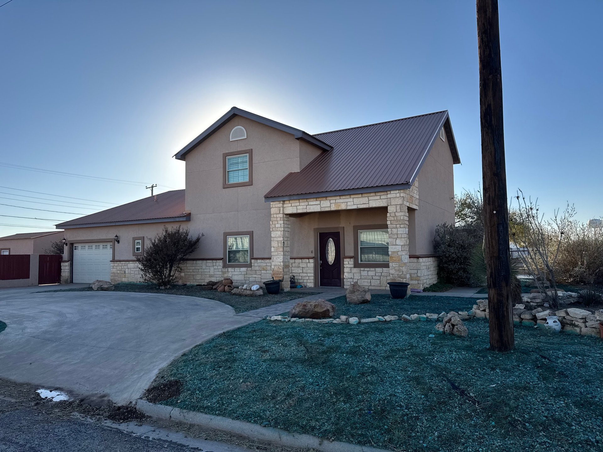 Modern single-story house with stone and tan exterior, metal roof, driveway, and landscaped front yard under clear blue sky